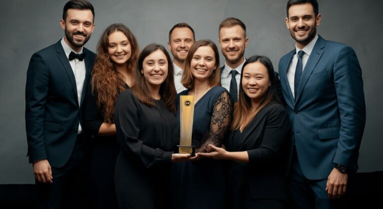 Diverse team in formal attire gathered around a table, jointly presenting a tall gold trophy with happy smiles, Global Recognition Awards.